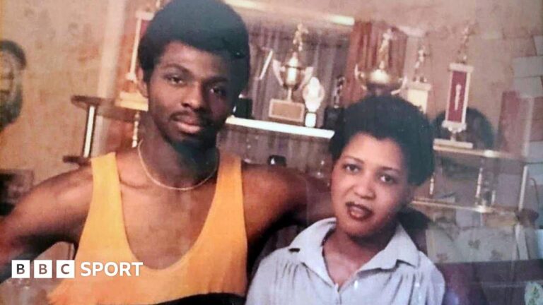 A young James Cook, wearing a yellow boxing vest, with his arm around his wife Carmen. There are various sport trophies behind them.