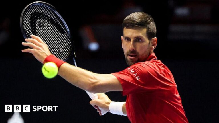 Novak Djokovic is wearing a red T-shirt and preparing to hit a ball with his tennis racquet