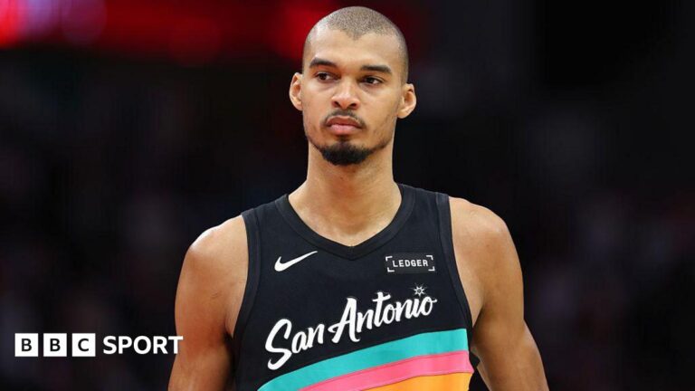 Victor Wembanyama wearing a vest that is black with blue, pink and orange stripes and the words San Antonio across the front