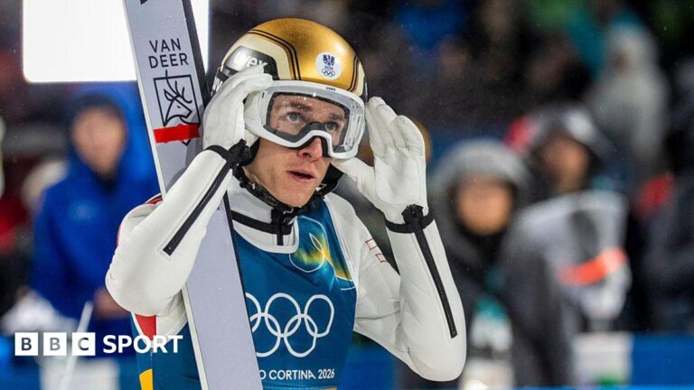 Daniel Tschofenig holds his hands up to his goggles and is wearing a gold helmet with a pair of skis on his shoulder
