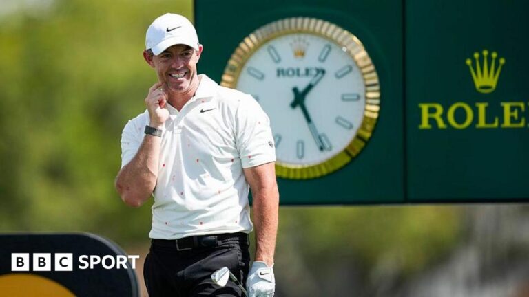 Rory McIlroy looks on after firing his tee-shot at the 14th hole of the Arnold Palmer Invitational. He is wearing a white cap, white Nike polo shirt and black trousers. He is holding his club in his gloved left hand. A large Rolex clock and advertising board can be seen behind him