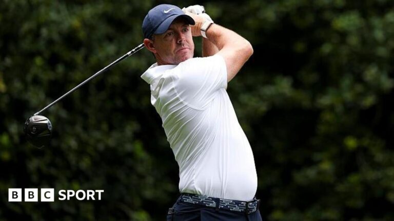 Rory McIlroy at the Truist Championship, wearing a white golf shirt and blue cap, swinging a metal wood