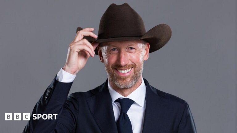 Graham Potter poses for the camera, wearing a suit while touching the brim of a cowboy hat