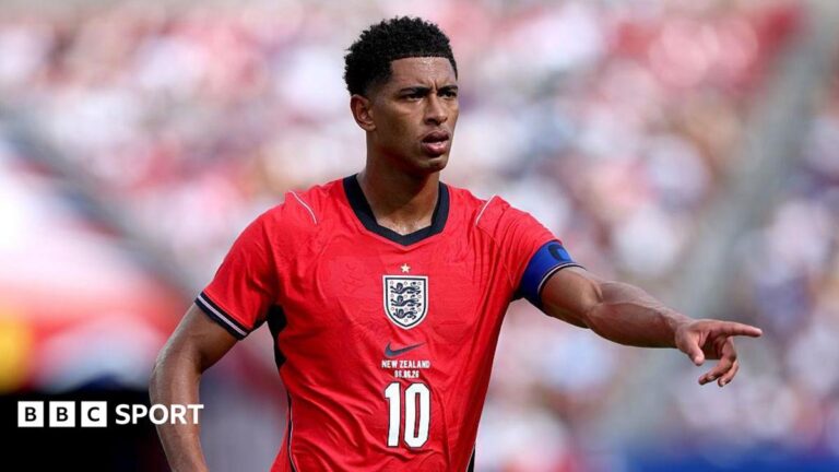 Jude Bellingham, wearing a red England shirt with a white number 10 in the centre beneath the badge, extends his left arm and points his index finger