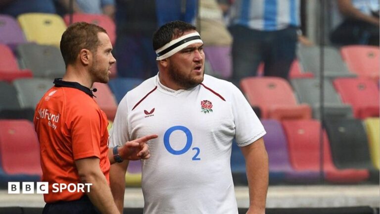 Referee Angus Gardner, wearing an orange top and dark blue shorts, raises a yellow card in his right hand