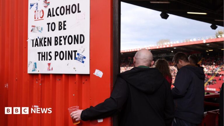 A football fan holds his pint of beer beneath a sign instructing that no alcohol should be taken beyond this point so he can continue to watch the match. The fan has his back to the camera and is wearing a dark-coloured jacket.