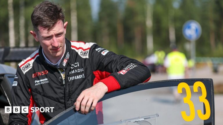 Elfyn Evans in black and red overalls leans on the open door of his number 33 rally car